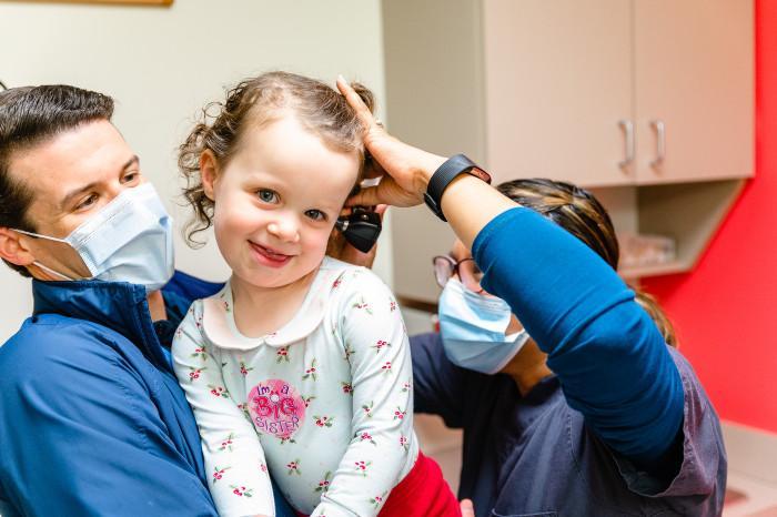 a smiling girl in her dads arms while a medical provider looks at her ear